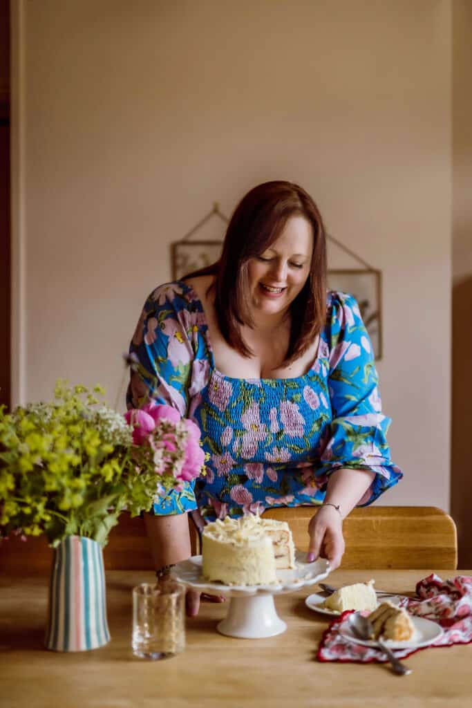 Terri Pugh is arranging a table. She is placing a cake on a stand, has some cake on plates already cut, and a vase contains some fresh wild flowers. She is wearing a light blue floaty top and is smiling.