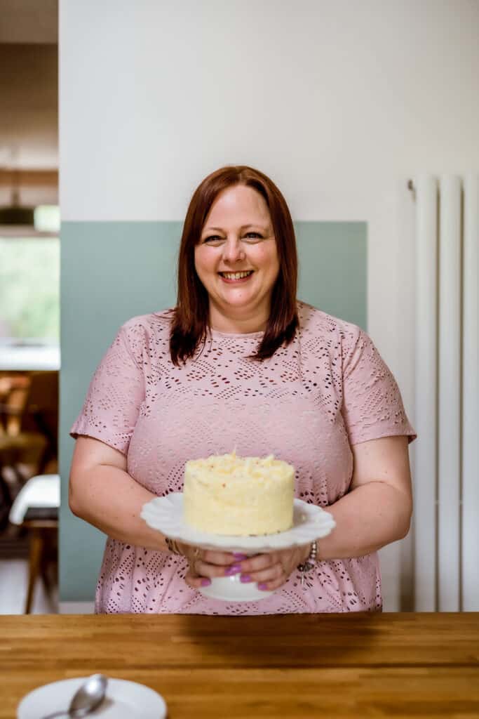 Terri Pugh is stood holding a cake stand which holds a lovely big white chocolate and raspberry cake. She is wearing a pink top, and looks very happy.