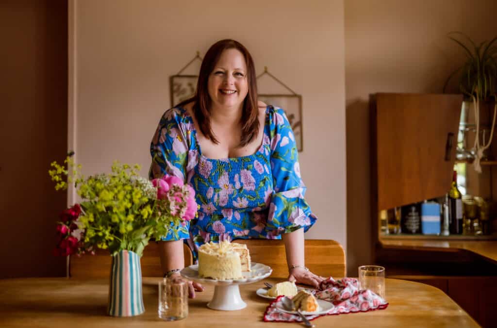Terri Pugh is leaning on a dining table which is laid out with cake on a stand, some cake on plates already cut, and a vase contains some fresh wild flowers. She is wearing a light blue floaty top and is smiling.