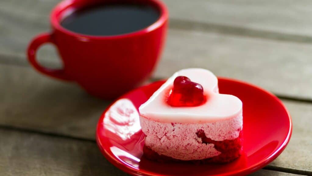 A sweet little heart cheesecake on a red plate, next to a coffee in a red cup